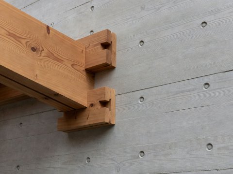 Close-up architectural study of interlocking cedar timber brackets meeting smooth board-formed concrete with tie-rod recesses
